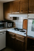 A cozy kitchen corner with wooden cabinets, a small stove with two black electric burners, and a black frying pan on one burner. A microwave and coffee maker are placed on the counter, and there are white tiled countertops with a backsplash. A cutting board is propped against the wall beside the stove.