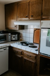 A cozy kitchen corner with wooden cabinets, a small stove with two black electric burners, and a black frying pan on one burner. A microwave and coffee maker are placed on the counter, and there are white tiled countertops with a backsplash. A cutting board is propped against the wall beside the stove.
