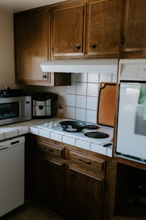 A cozy kitchen corner with wooden cabinets, a small stove with two black electric burners, and a black frying pan on one burner. A microwave and coffee maker are placed on the counter, and there are white tiled countertops with a backsplash. A cutting board is propped against the wall beside the stove.