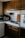 A cozy kitchen corner with wooden cabinets, a small stove with two black electric burners, and a black frying pan on one burner. A microwave and coffee maker are placed on the counter, and there are white tiled countertops with a backsplash. A cutting board is propped against the wall beside the stove.
