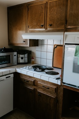 A cozy kitchen corner with wooden cabinets, a small stove with two black electric burners, and a black frying pan on one burner. A microwave and coffee maker are placed on the counter, and there are white tiled countertops with a backsplash. A cutting board is propped against the wall beside the stove.
