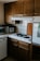 A cozy kitchen corner with wooden cabinets, a small stove with two black electric burners, and a black frying pan on one burner. A microwave and coffee maker are placed on the counter, and there are white tiled countertops with a backsplash. A cutting board is propped against the wall beside the stove.