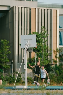 Two young people are playing basketball outdoors next to a modern building with vertical paneling. They are both focused and actively involved in the game as they attempt to reach the basket. The surrounding area includes greenery and well-maintained foliage, adding a natural touch to the urban setting.