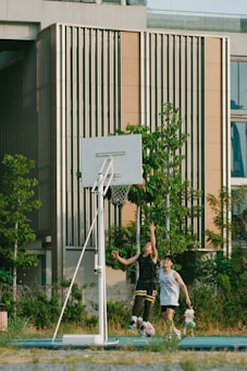 Two young people are playing basketball outdoors next to a modern building with vertical paneling. They are both focused and actively involved in the game as they attempt to reach the basket. The surrounding area includes greenery and well-maintained foliage, adding a natural touch to the urban setting.