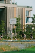 A group of people are playing basketball on an outdoor court near a modern building. The scene includes greenery and trees surrounding the court, and there is a clear sky suggesting a sunny day.