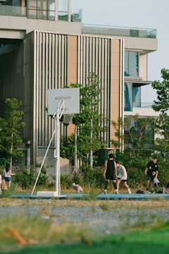 A group of people are playing basketball on an outdoor court near a modern building. The scene includes greenery and trees surrounding the court, and there is a clear sky suggesting a sunny day.