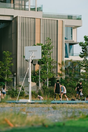 A group of people are playing basketball on an outdoor court near a modern building. The scene includes greenery and trees surrounding the court, and there is a clear sky suggesting a sunny day.