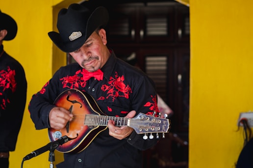 A musician wearing a black cowboy hat and a black shirt with red embroidery plays a string instrument. He is standing in front of a bright yellow wall, focusing intently on his music. A microphone is positioned in front of him, indicating he is performing.