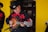 A musician wearing a black cowboy hat and a black shirt with red embroidery plays a string instrument. He is standing in front of a bright yellow wall, focusing intently on his music. A microphone is positioned in front of him, indicating he is performing.