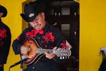 A musician wearing a black cowboy hat and a black shirt with red embroidery plays a string instrument. He is standing in front of a bright yellow wall, focusing intently on his music. A microphone is positioned in front of him, indicating he is performing.