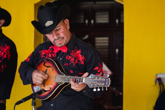 A musician wearing a black cowboy hat and a black shirt with red embroidery plays a string instrument. He is standing in front of a bright yellow wall, focusing intently on his music. A microphone is positioned in front of him, indicating he is performing.