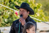 A man wearing a black cowboy hat and a black shirt holds a microphone. He appears to be speaking or singing, with greenery in the background indicating an outdoor setting. A young girl with dark hair stands in front of him, partially visible.
