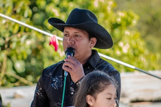 A man wearing a black cowboy hat and a black shirt holds a microphone. He appears to be speaking or singing, with greenery in the background indicating an outdoor setting. A young girl with dark hair stands in front of him, partially visible.