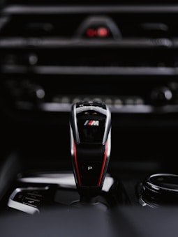 A sleek and modern car gear shifter with a black and red design, featuring the letter P and a logo prominently displayed. The background is blurred, highlighting the controls and buttons on the car's center console.