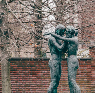 Two bronze sculptures depicting figures embracing stand in front of a brick wall. Behind them, tall trees with bare branches create a textured background, hinting at the season of autumn or winter. The earthy tones of the setting, along with a hint of green on the patina of the statues, establish a natural and serene environment.