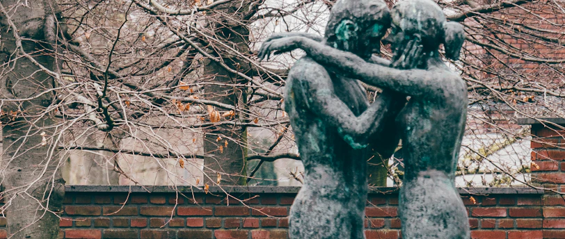 Two bronze sculptures depicting figures embracing stand in front of a brick wall. Behind them, tall trees with bare branches create a textured background, hinting at the season of autumn or winter. The earthy tones of the setting, along with a hint of green on the patina of the statues, establish a natural and serene environment.