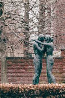 Two bronze sculptures depicting figures embracing stand in front of a brick wall. Behind them, tall trees with bare branches create a textured background, hinting at the season of autumn or winter. The earthy tones of the setting, along with a hint of green on the patina of the statues, establish a natural and serene environment.