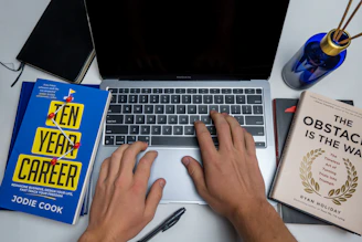 a person typing on a laptop next to a book