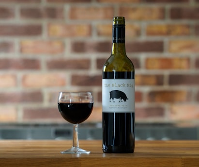 A bottle of red wine labeled 'The Black Pig' stands on a wooden surface beside a filled wine glass. The background consists of a brick wall with a mix of brown, red, and orange hues.