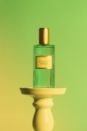 A perfume bottle with a gold cap and a transparent green body is placed on a cream-colored pedestal. The background features a gradient of green and yellow hues, creating a soft and elegant atmosphere.