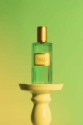 A perfume bottle with a gold cap and a transparent green body is placed on a cream-colored pedestal. The background features a gradient of green and yellow hues, creating a soft and elegant atmosphere.