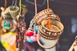 A collection of colorful handcrafted items hanging closely together. Wooden bracelets with intricate patterns, colorful pom-pom keychains, and other small craft items are visible. The background is blurred, suggesting a market or craft fair setting.