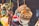 A collection of colorful handcrafted items hanging closely together. Wooden bracelets with intricate patterns, colorful pom-pom keychains, and other small craft items are visible. The background is blurred, suggesting a market or craft fair setting.