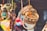 A collection of colorful handcrafted items hanging closely together. Wooden bracelets with intricate patterns, colorful pom-pom keychains, and other small craft items are visible. The background is blurred, suggesting a market or craft fair setting.