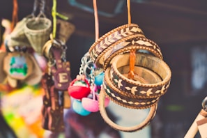 A collection of colorful handcrafted items hanging closely together. Wooden bracelets with intricate patterns, colorful pom-pom keychains, and other small craft items are visible. The background is blurred, suggesting a market or craft fair setting.