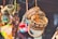 A collection of colorful handcrafted items hanging closely together. Wooden bracelets with intricate patterns, colorful pom-pom keychains, and other small craft items are visible. The background is blurred, suggesting a market or craft fair setting.