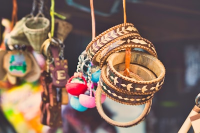 A collection of colorful handcrafted items hanging closely together. Wooden bracelets with intricate patterns, colorful pom-pom keychains, and other small craft items are visible. The background is blurred, suggesting a market or craft fair setting.