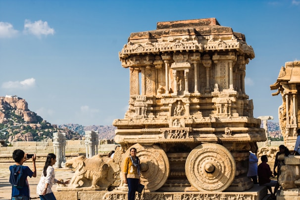 A large, intricately carved stone chariot situated in a historical site, surrounded by a few visitors. The chariot features elaborate decorations and sculptures depicting figures. In the background, there are rocky hills and a clear blue sky.