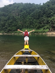 A man wearing a red life jacket stands with arms outstretched at the bow of a yellow boat. The boat is on a calm body of water surrounded by lush green forested hills under a cloudy sky.