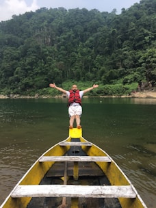 A man wearing a red life jacket stands with arms outstretched at the bow of a yellow boat. The boat is on a calm body of water surrounded by lush green forested hills under a cloudy sky.