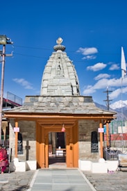A temple with intricate stone architecture and a wooden entrance. The structure features a tall spire with ornate carvings, set against a bright blue sky scattered with a few clouds. A person is seated to the side, dressed in traditional clothing. Mountains are visible in the background, adding to the scenic setting.