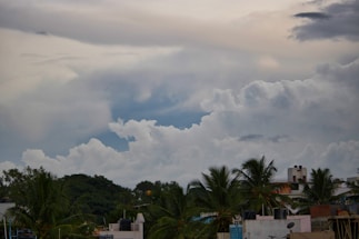 An urban landscape featuring a cluster of residential buildings with palm trees in the foreground. Above, the sky is filled with dramatic, thick clouds, varying from white to gray, suggesting a potentially stormy weather pattern. The tops of the buildings are dotted with water tanks and small antennas.