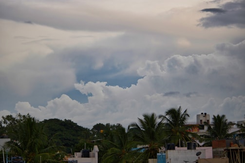An urban landscape featuring a cluster of residential buildings with palm trees in the foreground. Above, the sky is filled with dramatic, thick clouds, varying from white to gray, suggesting a potentially stormy weather pattern. The tops of the buildings are dotted with water tanks and small antennas.