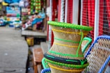 A vibrant market scene with colorful woven baskets prominently displayed in the foreground. The baskets are intricately designed with green and yellow plastic strands. The background shows a bustling market with various goods, stalls, and items along a narrow path.