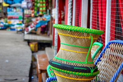 A vibrant market scene with colorful woven baskets prominently displayed in the foreground. The baskets are intricately designed with green and yellow plastic strands. The background shows a bustling market with various goods, stalls, and items along a narrow path.