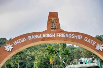 An arched gateway labeled 'India-Bangladesh Friendship Gate' with a national emblem at the top. The structure is flanked by decorative white flowers. Green trees and a partly cloudy sky form the background, contributing to a tranquil atmosphere.