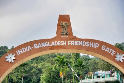 An arched gateway labeled 'India-Bangladesh Friendship Gate' with a national emblem at the top. The structure is flanked by decorative white flowers. Green trees and a partly cloudy sky form the background, contributing to a tranquil atmosphere.