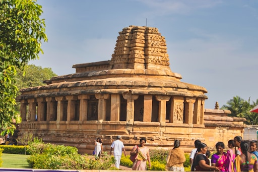 An ancient stone temple with intricate carvings and a stepped dome located in a landscaped area with lush greenery. Several people in colorful traditional clothing are gathered around the temple, engaging in various activities.