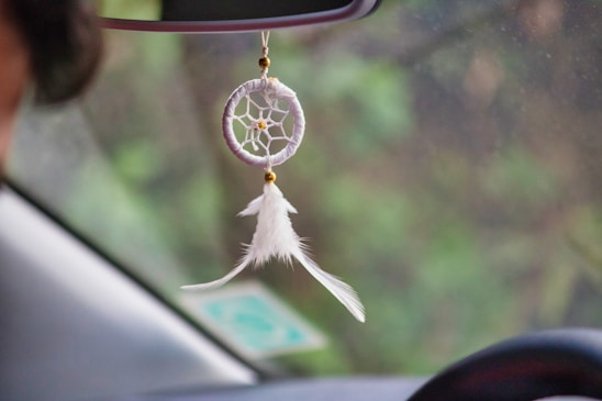 A dreamcatcher with white feathers and a beaded web hangs from a car's rearview mirror. The background shows a blurred view of greenery outside the car window.