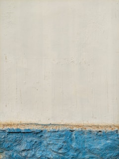 A textured wall with an upper portion painted in off-white and a lower portion in bright blue. The surface features visible brush strokes and signs of weathering. The separation between the colors is distinctly marked, suggesting a boundary between different materials or finishes.