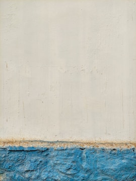 A textured wall with an upper portion painted in off-white and a lower portion in bright blue. The surface features visible brush strokes and signs of weathering. The separation between the colors is distinctly marked, suggesting a boundary between different materials or finishes.