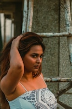 A woman with long hair wearing a patterned white top stands near a worn-out ladder structure. Her expression is confident and introspective, with one hand touching her hair.