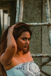 A woman with long hair wearing a patterned white top stands near a worn-out ladder structure. Her expression is confident and introspective, with one hand touching her hair.