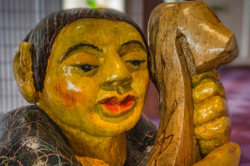 A detailed wooden sculpture depicting a human-like figure with yellow skin, large ears, and pronounced lips. The figure is holding a textured piece of wood close to its face, and the sculpture displays intricate carving details with a polished finish.