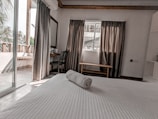 A neatly arranged bedroom with a white bed, a rolled towel on top, and natural light streaming in from the balcony. The room features grey curtains, a wooden chair, and a small table by the window. Palm trees are visible outside.