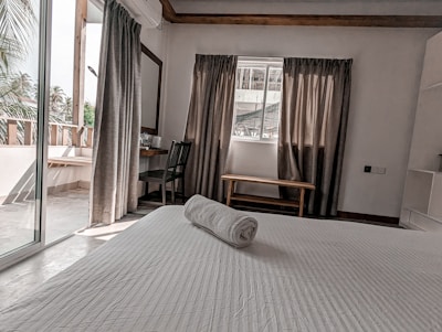 A neatly arranged bedroom with a white bed, a rolled towel on top, and natural light streaming in from the balcony. The room features grey curtains, a wooden chair, and a small table by the window. Palm trees are visible outside.